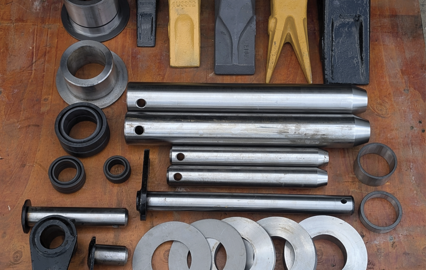 FLECO replacement parts including bucket teeth, cutting edges, pins, bushings, and shims laid out on a workbench